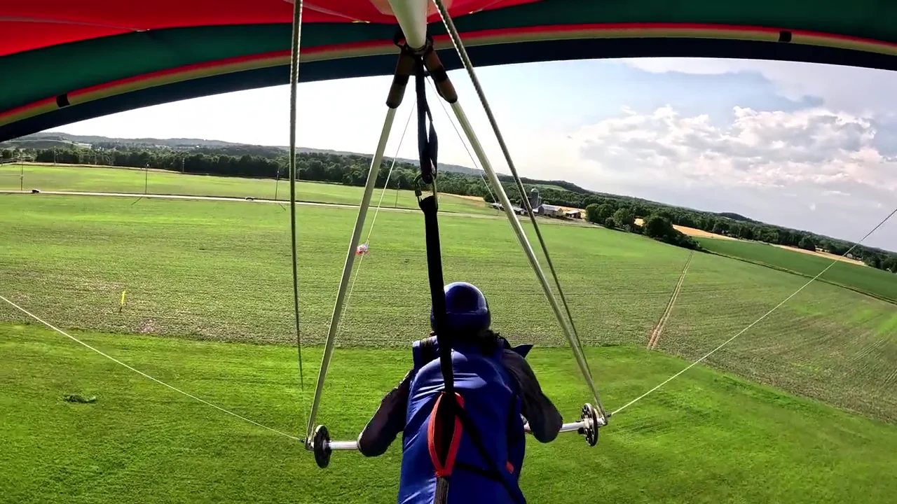 A hang-glider pilot beneath a colorful wing, flying over green fields at Padgham Hill